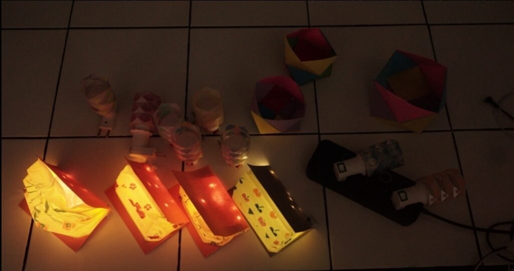 Hasil karya Origami Lamp buatan ibu-ibu PKK RT 26 yang memancarkan pendar cahaya estetik berpola geometris saat sesi pengujian lampu (lighting test) di ruangan yang diredupkan.
