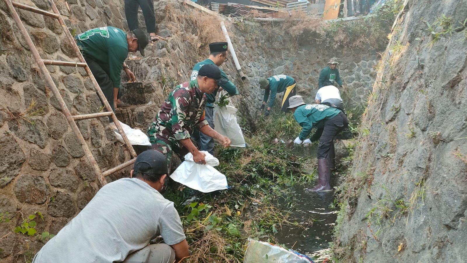 Babinsa Kelurahan Bareng Koramil 0833-01/Klojen, Serka Gatot (berseragam loreng TNI), membaur bahu-membahu bersama puluhan warga membersihkan tumpukan sampah dan sedimen saluran air dalam pelaksanaan GASS di Kelurahan Bareng, Senin (14/09).