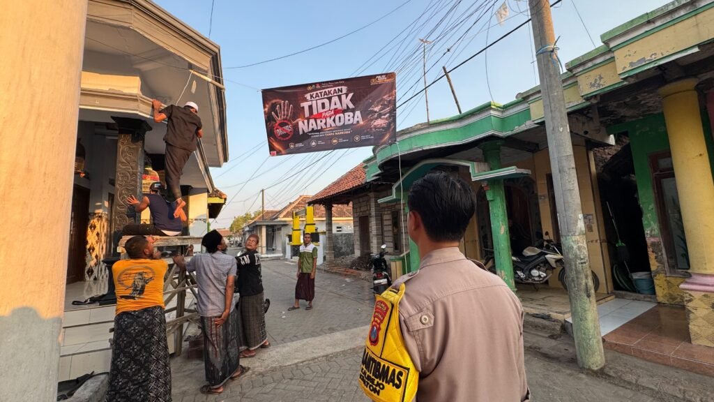 Suasana gotong royong saat warga dan aparat kepolisian memasang spanduk antinarkoba di lokasi strategis pemukiman warga.